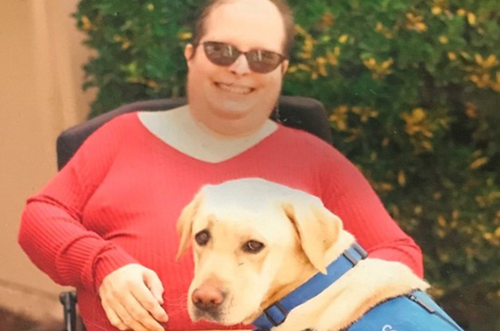 A person in a red shirt and sunglasses sits in a wheelchair, smiling, with a yellow service dog wearing a blue 