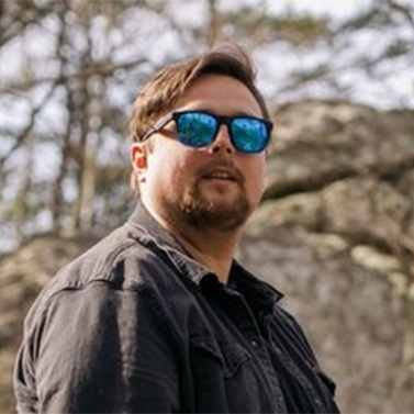 A man with short brown hair and a beard wears reflective blue sunglasses and a black jacket, standing outdoors with rocks and trees in the background.