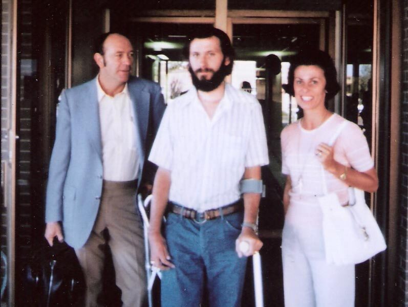 Three adults stand together outside a building entrance. The man in the center has a beard and uses crutches, while the other man and a woman stand on either side of him, smiling at the camera.