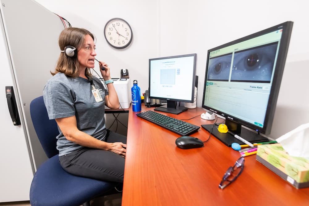 A woman wearing a headset sits at a desk with dual monitors. One screen shows images of eyes while the other displays data. A clock and webcam are on the wall, and various office items are scattered as she speaks into the headset.