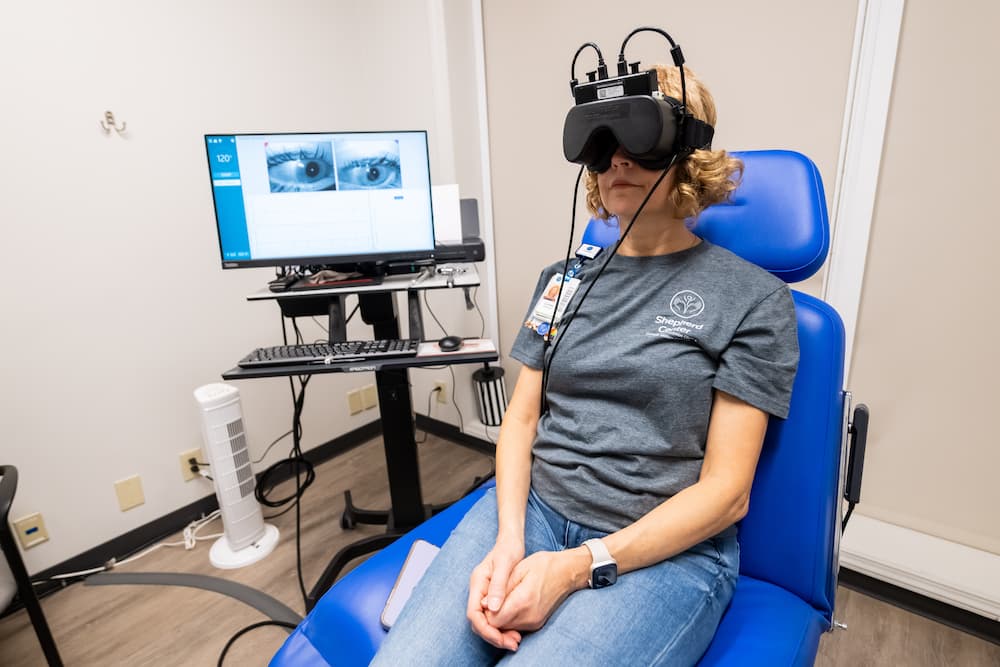 A person wearing virtual reality goggles sits on a blue chair in a testing room. A nearby monitor displays eye images and data, indicating they're part of an experiment.