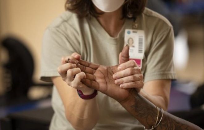 A healthcare worker wearing a face mask and ID badge holds and examines the hand of another person. The worker appears to be assessing or providing therapy, with focus on the thumb and fingers.