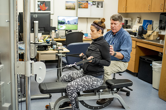 A woman in athletic wear performs resistance exercises on a machine while seated. A man in a blue shirt stands behind her, offering guidance. The room has offices and computers in the background.