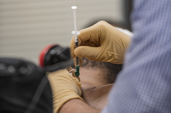 A person wearing a white hat and attached to monitoring electrodes is seated, with their back exposed. Another person, wearing gloves, prepares to inject or draw from the back with a syringe. A red device is partially visible on the right.