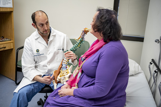 A doctor in a white coat shows a spine model to a seated woman in a purple outfit. They are in a medical office, discussing spine health. The doctor points at the model while the woman attentively listens.