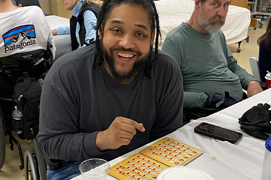 A smiling man sits at a table playing bingo, pointing at his bingo card. Another man with a beard sits beside him, looking at the camera. Both are in wheelchairs, and a drink and phone are on the table.