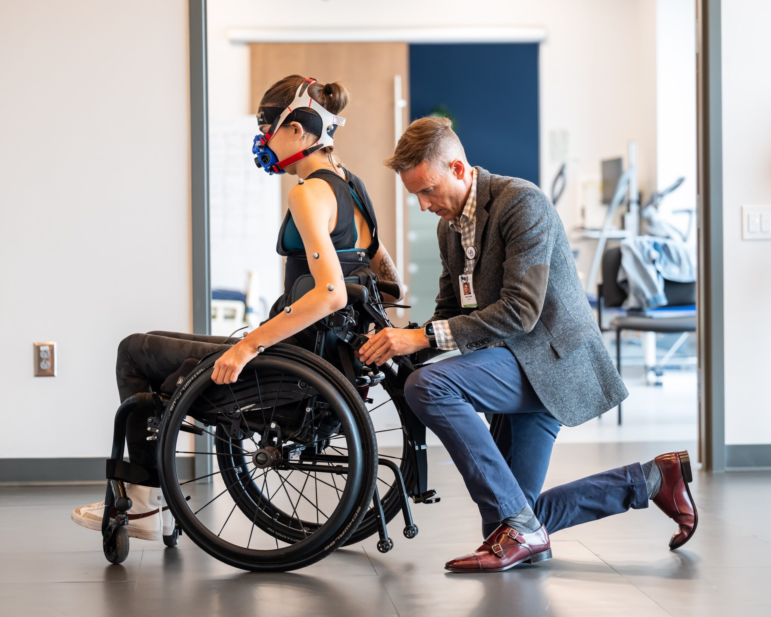 A woman in a wheelchair wears fitness sensors and a breathing mask while a man in a blazer kneels beside her, adjusting equipment on her wheelchair in a modern, clinical setting.