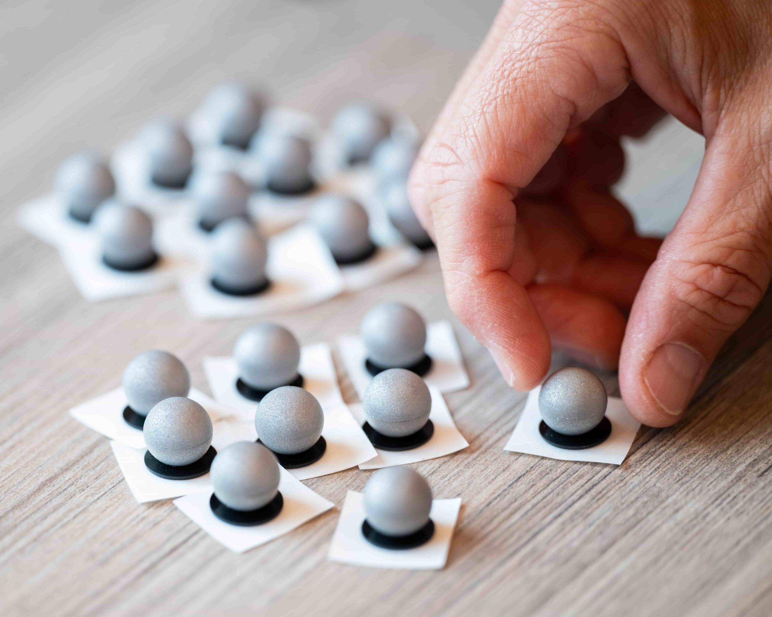 A hand picks up a small, round, metallic gray object from a group of similar objects, each placed on a square white paper with a black circle, arranged on a wooden surface.