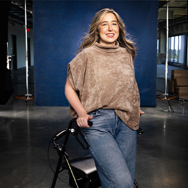 A woman with long, wavy hair smiles while standing and holding a walker. She wears a loose tan top and jeans, and stands in a modern indoor space with a blue backdrop behind her.