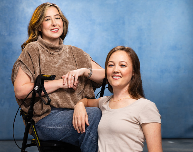 Two women pose and smile in front of a blue backdrop. One woman sits in a wheelchair wearing a brown top and jeans, while the other kneels beside her, dressed in a light beige shirt.