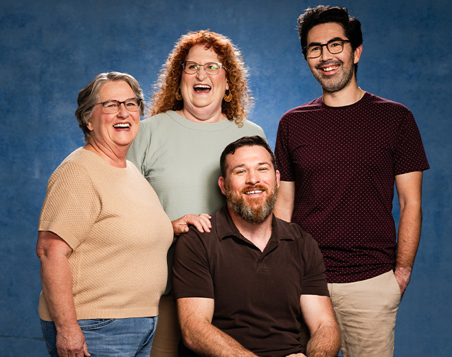 Four adults, three standing and one seated, smile and pose together against a blue backdrop. The group includes two women and two men, all wearing casual clothing and eyeglasses, appearing cheerful and friendly.