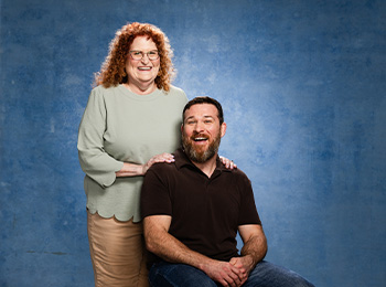 A woman with curly red hair stands behind a seated bearded man. She rests her hands on his shoulders. Both are smiling in front of a blue textured background.