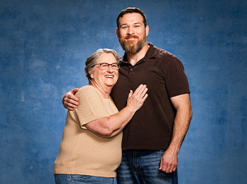 An older woman with short gray hair and glasses smiles while hugging a bearded man in a dark polo shirt against a blue background. Both look happy and relaxed.