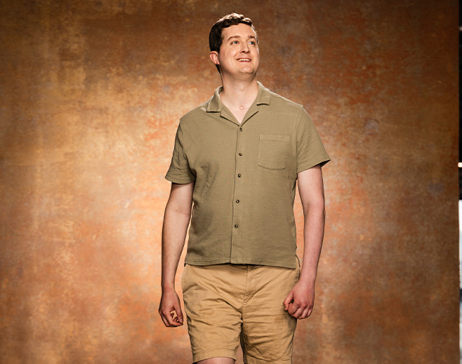A man wearing a short-sleeve, olive-green button-up shirt and tan shorts stands smiling in front of a textured brownish background.