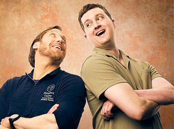 Two men stand back to back with their arms crossed, smiling and looking at each other against a warm, textured background. One wears a navy zip-up and the other a khaki polo shirt.