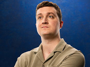 A young man with short brown hair wearing a beige collared shirt looks up confidently against a solid blue background.