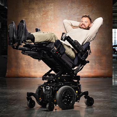 A man smiles and reclines comfortably in a high-tech electric wheelchair with his legs raised and hands behind his head, set against a brown studio backdrop.