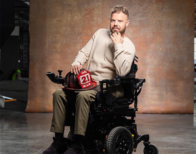 A man sits in a motorized wheelchair, wearing a beige sweater and olive pants, holding a firefighter’s helmet marked 