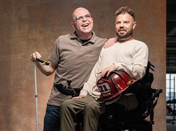 Two men pose together, smiling. One stands holding a white cane and has his arm around the other, who is seated in a power wheelchair and holding a firefighter helmet with the number 21 on it.