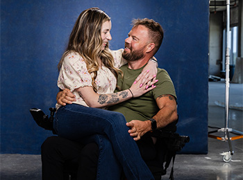 A woman sits on a man's lap, both smiling and looking at each other affectionately. The man has a beard and wears a green shirt, while the woman has long hair and wears a floral blouse and jeans. They are indoors with a blue background.
