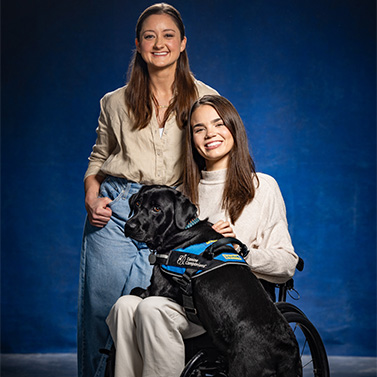 Two women pose together in front of a blue background. One woman sits in a wheelchair with a service dog in a harness, while the other stands beside her, smiling.
