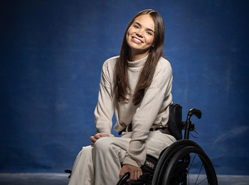 A young woman with long brown hair, wearing a light sweater and pants, smiles while sitting in a wheelchair against a blue background.