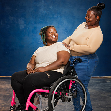 Two women smile warmly at each other; one is seated in a pink wheelchair, while the other stands beside her, resting a hand on her shoulder, both wearing light-colored tops against a blue backdrop.