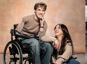 A young man in a wheelchair and a young woman kneeling beside him both smile warmly at the camera against a neutral, textured background. They are wearing casual beige sweaters and blue jeans.