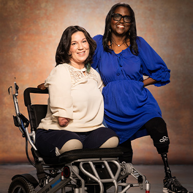 Two women with limb differences smile at the camera. One woman sits in a wheelchair and wears a cream blouse, while the other stands beside her in a blue dress with a prosthetic leg. They pose against a warm, neutral backdrop.