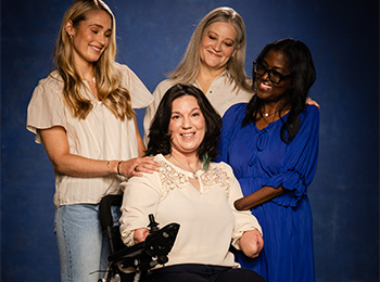 Four women pose together smiling against a dark blue backdrop. The woman in front uses a wheelchair and has limb differences; the three women behind her rest their hands on her shoulders, showing support and friendship.