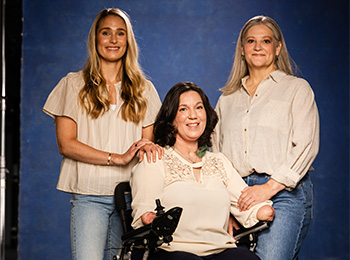 Three women pose together in front of a blue backdrop. One woman sits in a wheelchair and has limb differences, while the other two women stand on either side of her with their hands on her shoulders. All are smiling.