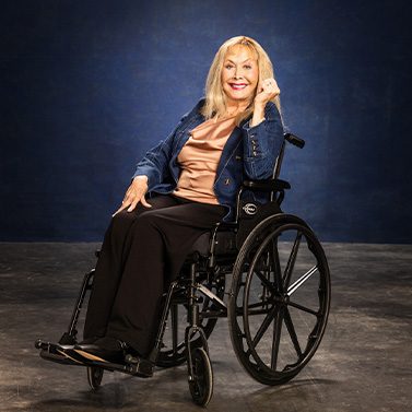 A smiling older woman with long blond hair sits in a wheelchair against a dark blue background. She is wearing a blue jacket, tan top, and black pants, and looks confidently at the camera.