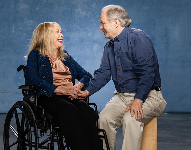 An older woman in a wheelchair smiles and holds hands with an older man sitting on a stool. Both are dressed casually and look happy against a blue background.