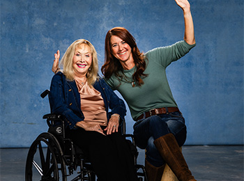 Two women smiling and waving at the camera; one is seated in a wheelchair, wearing a pink blouse and dark pants, while the other, in a green sweater and jeans, sits beside her on a stool. Blue background.
