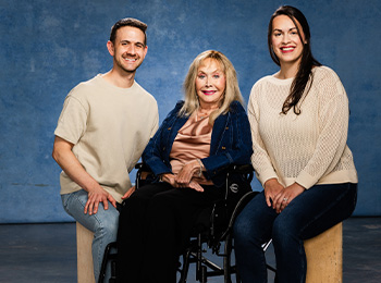 Three people pose together in front of a blue backdrop: a smiling older woman in a wheelchair sits between a man and a woman, who are also smiling and seated on wooden stools.