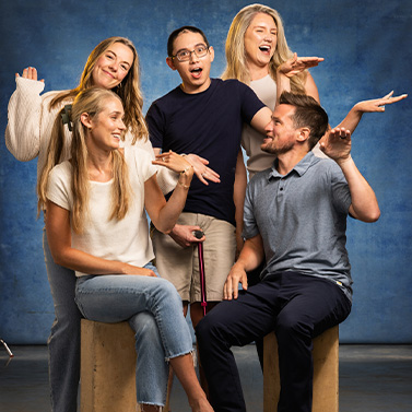 Five young adults pose playfully against a blue backdrop, smiling and holding their hands up in a fun gesture. Two are seated on stools while three stand behind them, all appearing cheerful and energetic.