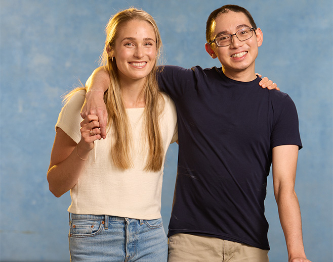 Two young adults, one woman and one man, stand against a blue background. The man has his arm around the woman’s shoulders, and both are smiling at the camera. They appear relaxed and friendly.