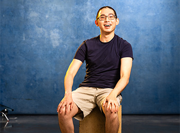 A man with glasses, wearing a dark t-shirt and beige shorts, sits on a wooden box and smiles in front of a blue mottled background.