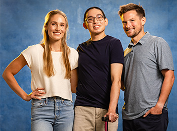 Three people stand together smiling in front of a blue background. The woman on the left wears a white shirt and jeans, the man in the middle wears glasses and holds a cane, and the man on the right wears a gray polo shirt.