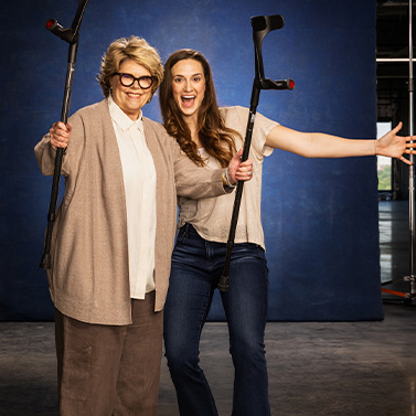 Two smiling women pose together. The older woman wears glasses and a beige outfit, while the younger woman holds black forearm crutches in both hands and spreads her arms wide, standing in front of a blue background.