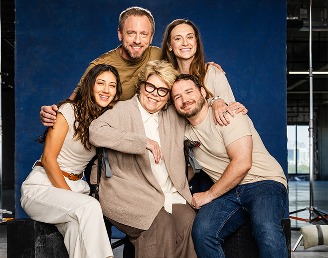 Five people pose together, smiling warmly. Three are seated, including an older woman in the center, with two younger men and two women, one standing behind and one seated, all casually dressed against a blue studio backdrop.
