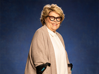 A smiling older woman with short, wavy blonde hair and black glasses stands against a blue background, wearing a light blouse and a beige shawl draped over her shoulders.