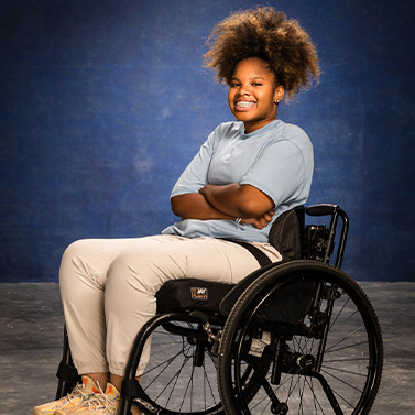 A young person with curly hair sits smiling in a wheelchair against a blue background, wearing a light blue shirt, light pants, and sneakers, with arms crossed and a confident expression.