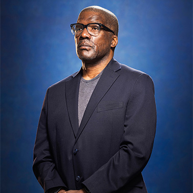 A man wearing glasses, a dark blazer, and a gray shirt stands with his hands folded, looking slightly upward against a blue gradient background.