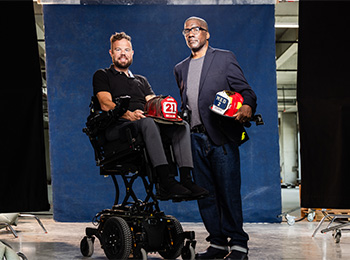 Two men pose for a photo in a studio. One man sits in a power wheelchair holding a firefighter helmet, while the other stands beside him, also holding a helmet. They are in front of a blue backdrop.