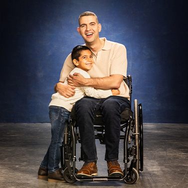 A man in a wheelchair smiles and embraces a young boy who is standing next to him, both appearing happy against a dark blue background.