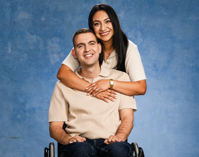 A smiling man in a wheelchair sits in front of a blue background while a woman stands behind him, hugging him around the shoulders. Both are wearing light-colored shirts and look happy.