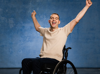A smiling man in a wheelchair raises both arms in celebration against a blue background.