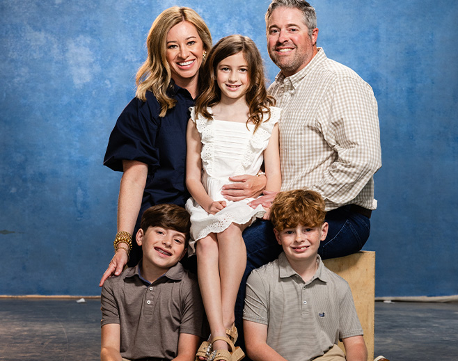 A family of five poses and smiles in front of a blue background. A woman and man sit behind a young girl in a white dress, with two young boys in polo shirts sitting in front of them on the floor.