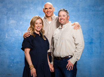 Three adults pose together in front of a blue backdrop. Two men stand side by side, one with his hand on the other’s shoulder, while a woman stands next to them, smiling at the camera.